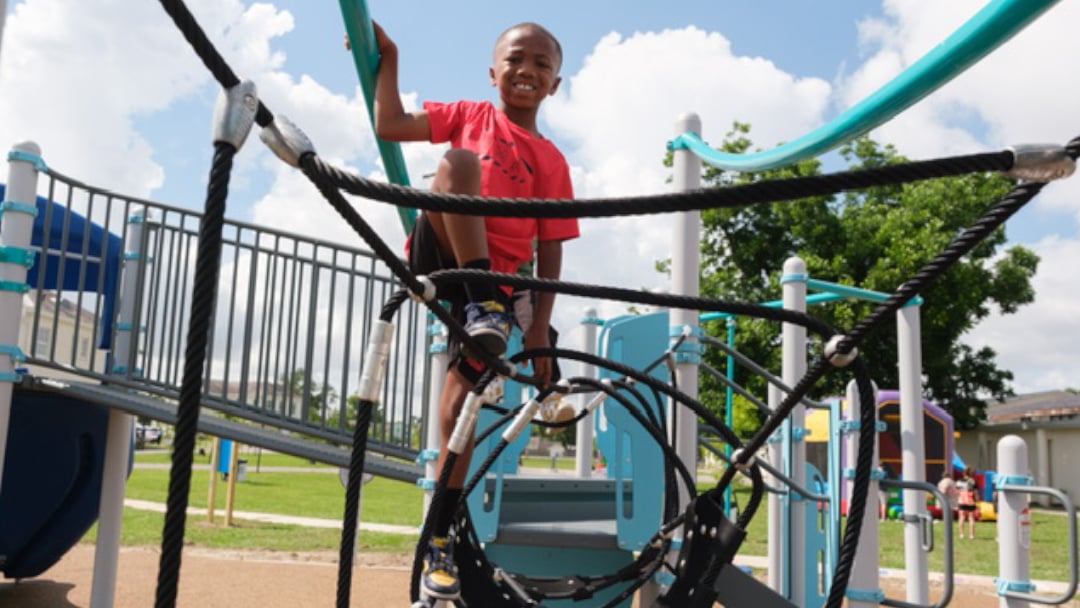 A young boy in a red shirt climbs a rope structure on the new KaBoom! playground, smiling and enjoying the outdoor play equipment under a clear blue sky.