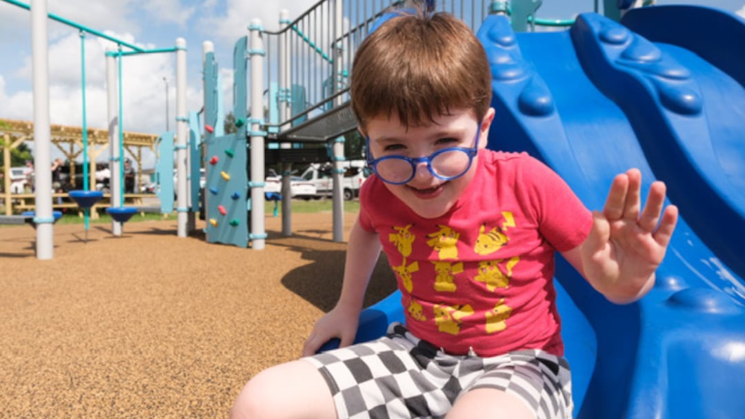 A joyful child in red shirt and checkered shorts sits at the top of a blue playground slide, waving with a bright smile, surrounded by colorful play equipment in the new KaBoom playground.