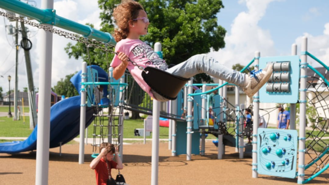 A young girl swings happily on a playground swing set, surrounded by outdoor play equipment in a park setting.