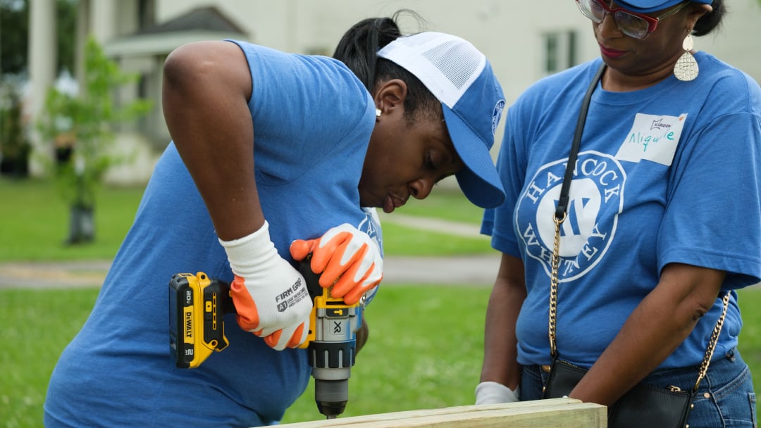 Two Hancock Whitney volunteers in blue shirts use a power drill to assemble a wooden structure, working together to build the new KaBoom! playground components.