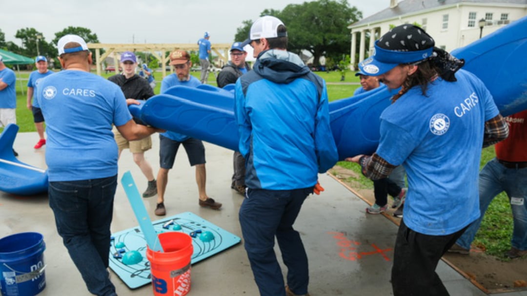 Several Hancock Whitney volunteers in blue shirts and jackets carry large blue KaBoom! playground pieces across a construction site, with buckets and equipment nearby.