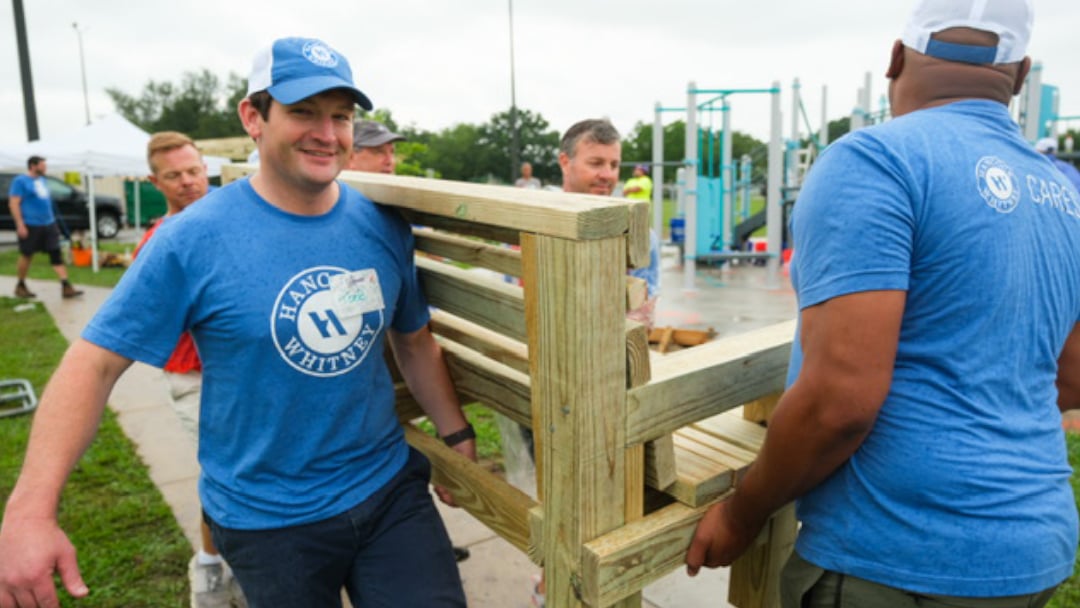 Two Hancock Whitney volunteers in blue shirts carry a wooden bench together at a KaBoom! playground construction site, with other volunteers and playground equipment in the background.